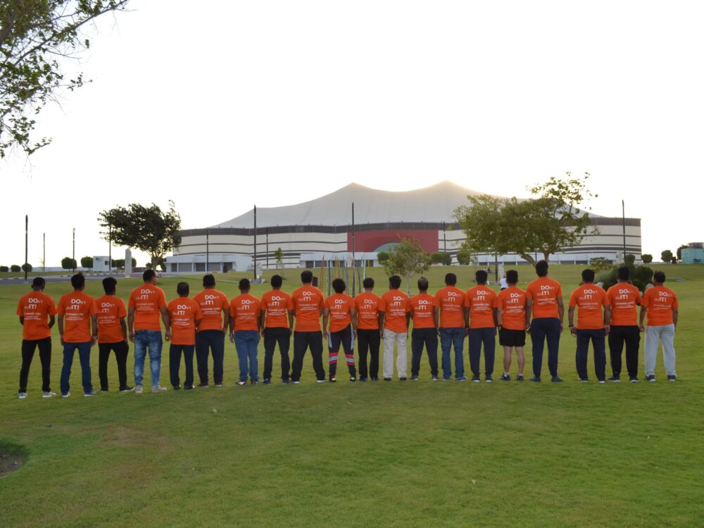 Group of people standing in a line on grass wearing matching orange shirts with text on the back facing away from the camera