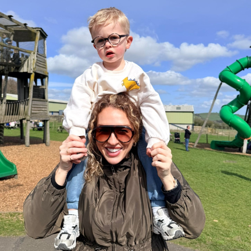Portrait pic of Tegan with her young son on her shoulders, she is standing at a playground, smiling happily into the camera
