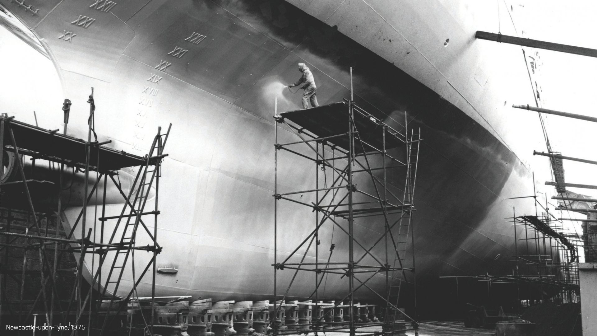 blck & white photo of a large ship in the dock being repainted in 1975. A worker is working with a spray gun on a high scaffold, without any railing on the upper deck